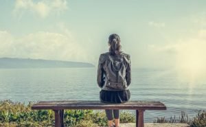 A lonely woman sits on a bench facing a body of water.